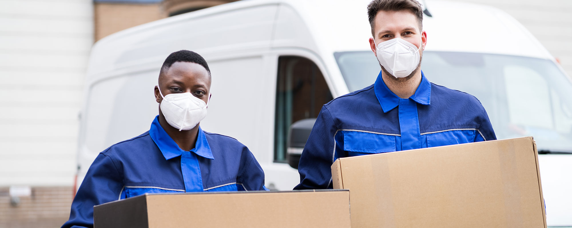 two adult males with masks over their noses and mouths holding moving boxes with their white moving vehicle in the background